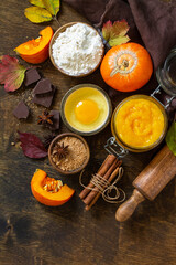 Seasonal food background - ingredients for autumn baking (pumpkin puree, eggs, flour, chocolate, sugar and spices) on a wooden table. Top view flat lay background. Copy space.