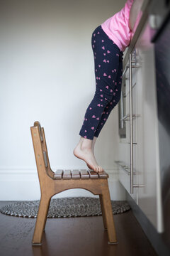 A Young Child Girl Standing On A Small Chair Stretching To Reach The Tap In A Kitchen 