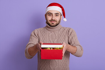 Bearded guy in santa hat and beige sweater brings gifts in red box, posing isolated over lilac background, looks at camera, young handsome male with present.