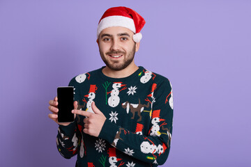 Smiling young Santa European man wearing sweater with snowmen and Christmas hat, holds mobile phone with empty blank screen isolated on purple background. New Year celebration.