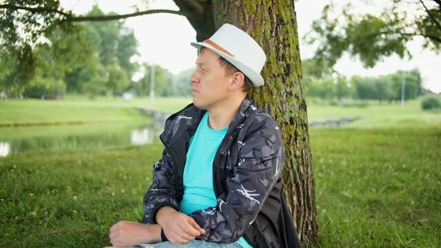 Young man in a white hat sits on a tree, looks into the distance, tracking camera