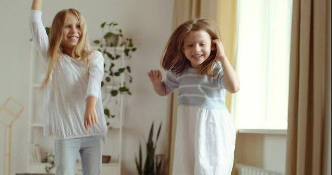 Two little caucasian blonde girls jumping together at home on sofa or bed, sisters spend time having fun, two children laughing smiling playing in room during quarantine isolation, childhood concept