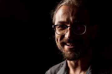 Dark studio portrait of a middle-aged man with beard in glasses