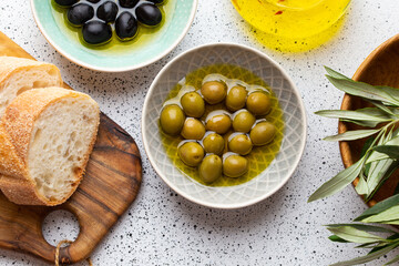 Green and black olives with olive oil in a glass bottle, olive tree sprigs and cut fresh ciabatta bread on wooden cutting board. White rustic background, mediterranean food concept, top view