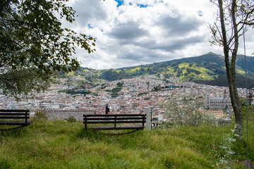 bench in the mountain looking at a beautiful historic city surrounded by grass, branches and trees with white clouds 