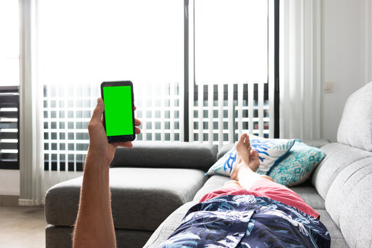 A man lying on a sofa holds a mobile with a green screen with one hand