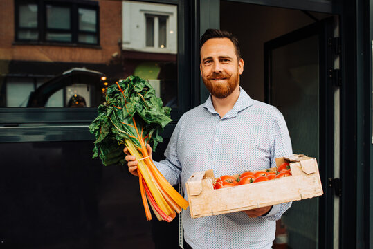 Delivery man holding box of tomatoes and greens at restaurant entrance