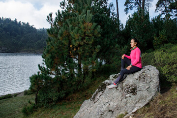 Naklejka na ściany i meble Mujer sentada en una roca a la orilla de un lago con arboles al fondo