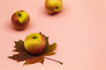 In the horizontal photo from the side of the pink background are whole apples and a sheet of maple