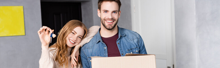 panoramic shot of joyful woman holding keys near boyfriend with carton box, moving concept