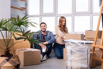 man sitting on floor with cup while woman unpacking carton boxes in new home