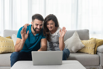 Young couple sitting in their living room with laptop waving to their marital therapist after...