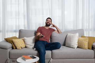Young casual man sitting on the sofa at his home with remote control in his hand relaxing with...