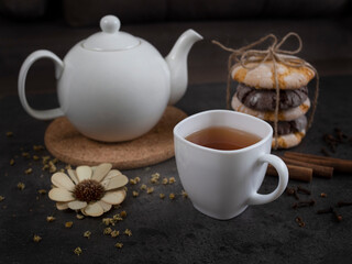 Fototapeta na wymiar White teapot with a cup of black tea, orange and chocolate cookies, dried flower, several sticks of cinnamon on the brown wooden background. Focus on a cup. Close up. 