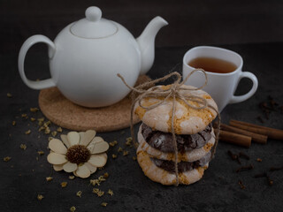 White teapot with a cup of black tea, orange and chocolate cookies, dried flower, several sticks of cinnamon on the brown wooden background. Focus on cookies. Close up. 