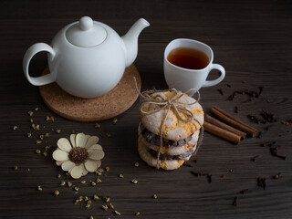 Fototapeta na wymiar White teapot with a cup of black tea, orange and chocolate cookies, dried flower, several sticks of cinnamon on the brown wooden background. Focus on cookies.