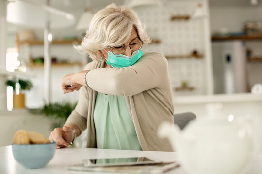 Senior woman with face mask sneezing into elbow at home. - Powered by Adobe