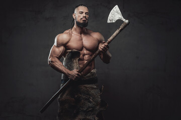 Northen bearded barbarian with huge biceps and dreadlock posing while holding axe on his hands in...