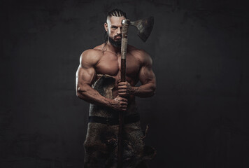 Ancient muscular viking staying with naked torso and dreadlocks holding his axe hiding half of his face in dark background.