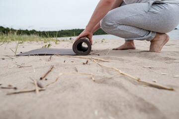 Man unfolds a yoga mat on a sandy beach near a lake, he is about to do exercises outdoors on a...