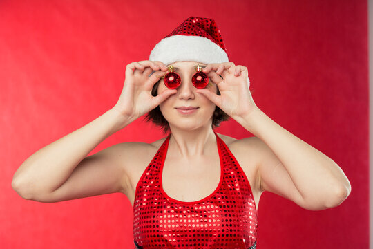 Portrait a girl in a Santa Claus hat is holding red Christmas balls in front of her eyes and smiling