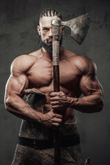 Bodybuilder in fashion of furious viking posing holding axe near face with naked torso in dark background.