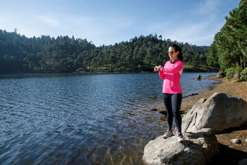 Mujer corredora usando su reloj inteligente despu&eacute;s de correr, sentada en la orilla del  lago sobre una piedra