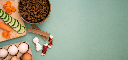 Dry pet food with natural ingredients. Raw meat, vegetables cucumber and carrot near eggs on green background with copy space