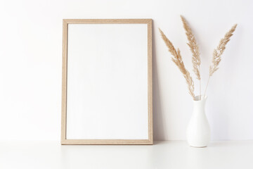Empty wooden frame stands on white table with dry grass in vase. Mockup poster frame close up in...