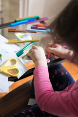 Looking down at a young girl child seated at her study desk learning school from home with ipad tablet education program colored colorful textas scissors glue