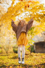 A little girl in a yellow dress and a brown sweater jumps in the autumn Park. A girl with red hair and a yellow dress jumps in an autumn Park. Autumn photo shoot of a girl with red hair.
