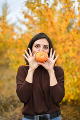 A woman and a pumpkin on the background of an autumn forest.  The frightened girl holds a small pumpkin. Halloween. Vertical photo