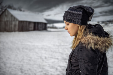 Side view of a beautiful blonde woman looking down, wearing a black winter coat and a Beanie hat, standing alone in a snowy winter rural landscape with a cloudy sky