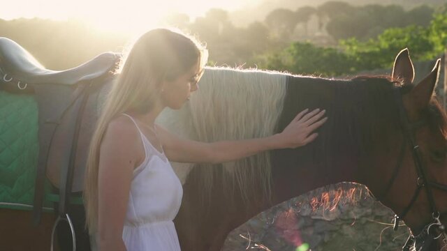 Young beautiful woman caressing a brown and white cherokee horse at sunset. Female wearing white greek classical dress touching a horse in the forest with nice sunset light with dreamy mood.
