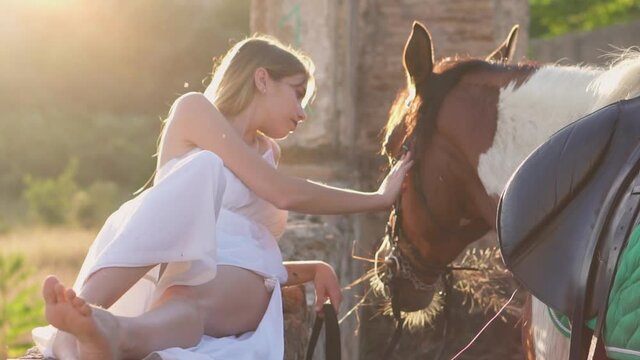 Woman touching and caressing a horse while it is grazing grass in the forest and mountain. Beautiful sunset and light. Model wearing roman white dress or toga enjoying with a horse