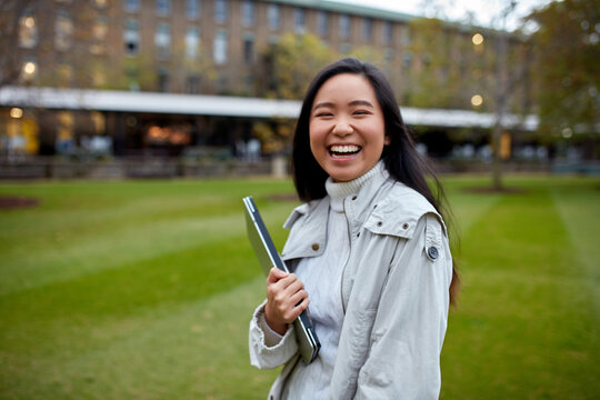 Young Asian student holding her laptop on lawn at university campus