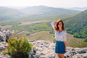 A beautiful woman stands on a high cliff, enjoying the view of the mountain range and forest