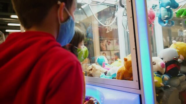 Masked boys due to viral quarantine playing games of chance in public area. Flashing LED lights and sound of crane toy machine as birthday gift. Gambling elated excited mood.
