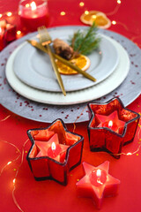Traditional Christmas table place setting. Golden cutlery, gray plates, red candles, glitters, spruce branche and pine coner. Holidays red background.