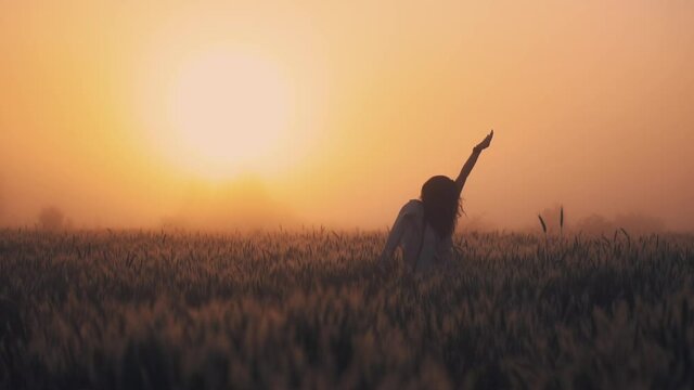 A girl stands in the middle of a wheat field, waving at dawn in a thick fog
