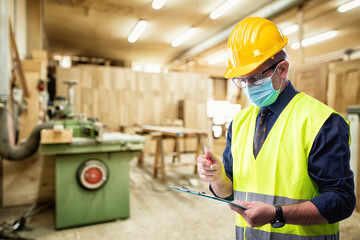 Engineer in carpentry workshop wears surgical mask to prevent Coronavirus spread, holds pen and notebook in hand. Preventing Pandemic Covid-19 at the workplace.