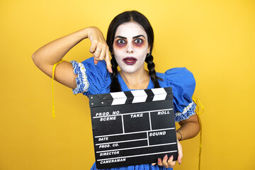 woman wearing a scary doll halloween costume over yellow background holding clapperboard very happy...
