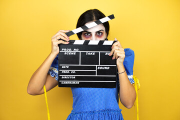 woman wearing a scary doll halloween costume over yellow background holding clapperboard very happy...