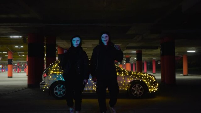People with luminous purge masks are holding a baseball bat and katana. Murderers at night in the parking lot against the backdrop of a car with a garland.