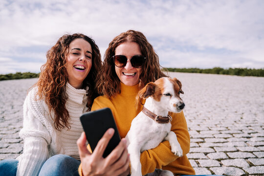 Two beautiful women sitting with a little dog in a park of Madrid and trying to take a photo with the phone. Technology lifestyle with dogs