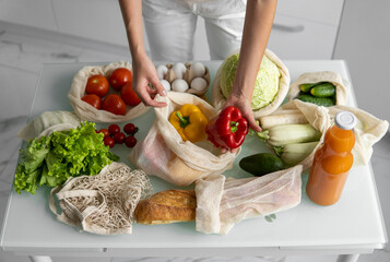 Woman's hand, holding a reusable grocery bag with vegetables on a kitchen at home and takes pepper out. Zero waste and plastic free concept. Mesh cotton shopper with vegetables. Ecology.