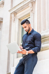 Young East Indian American Businessman with beard working in New York City, wearing black shirt, holding laptop computer, standing outside old style office building, looking down, reading, typing..