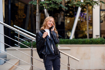 Cheerful beautiful student girl speaks on smart phone at stairs on the street, enjoying walk outdoors.