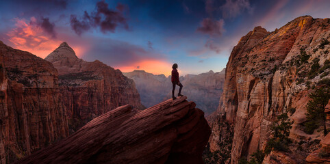 Adventurous Woman at the edge of a cliff is looking at a beautiful landscape view in the Canyon during a vibrant sunset. Taken in Zion National Park, Utah, United States. Sky Composite Panorama - obrazy, fototapety, plakaty