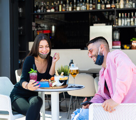 smiling couple of a happy man an a woman with face mask having a drink in a cafè bar looking at the smartphone. concept about the joy and fun to spend time together drinking beer in the new normality